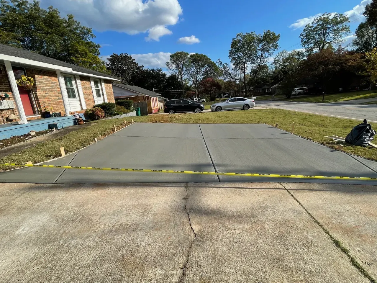 Rural driveway blocked by a yellow tape barrier, with houses, cars, and trees under a blue sky.
