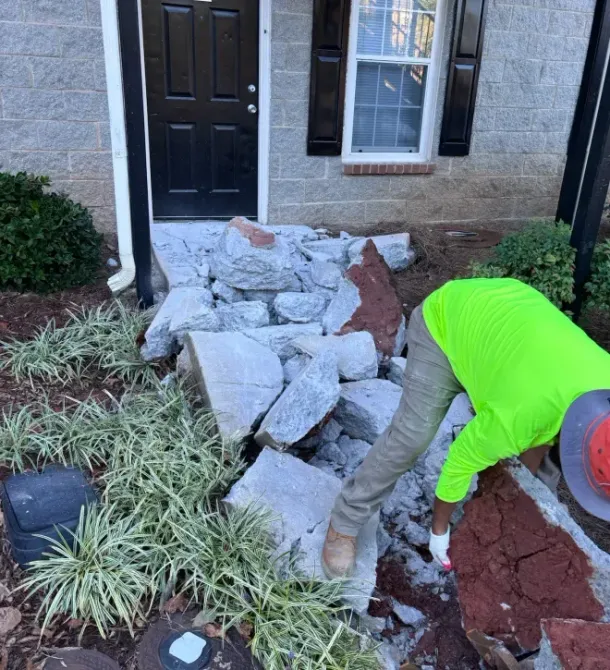 A worker in a neon green shirt lifts a large rock from a pile of stone debris in front of a building entrance.