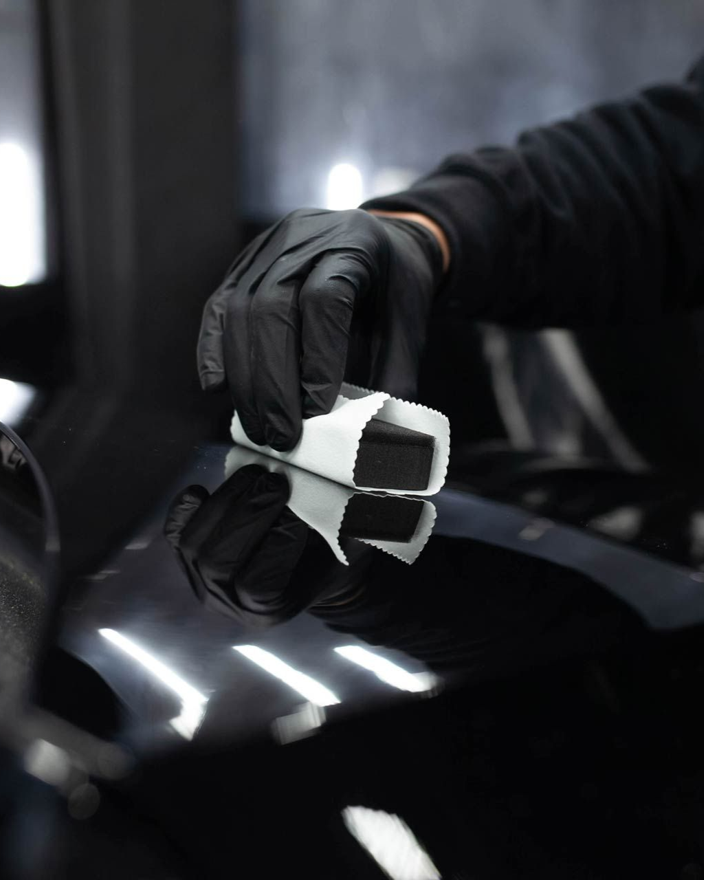 Black-gloved hand applying product to a shiny, black car surface with an applicator sponge.
