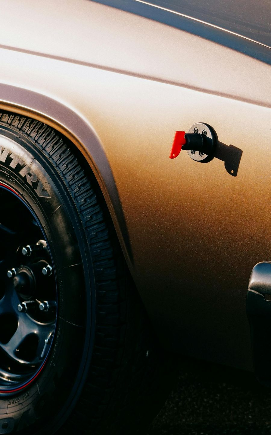 Close-up of a vintage brown car's wheel and side panel, with a black fuel door and red release lever.