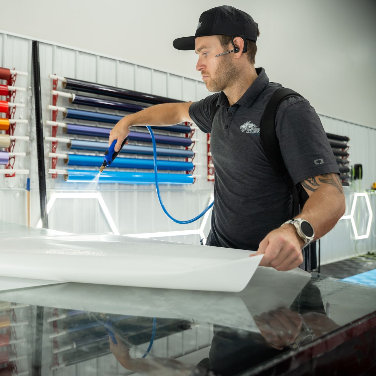 Man spraying water on a white sheet in a workshop, rolls of colored material on the wall.