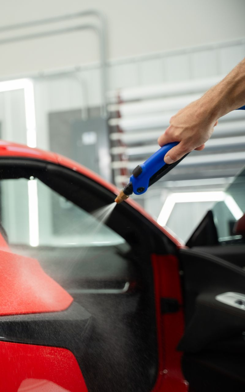 Person cleaning a red car with a blue spray nozzle; interior shot.