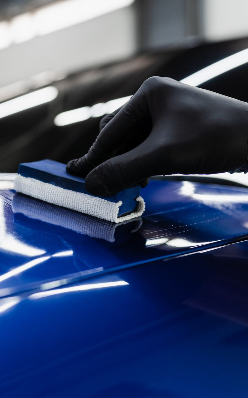 Hand in black glove applying blue coating with applicator to a blue car's surface.