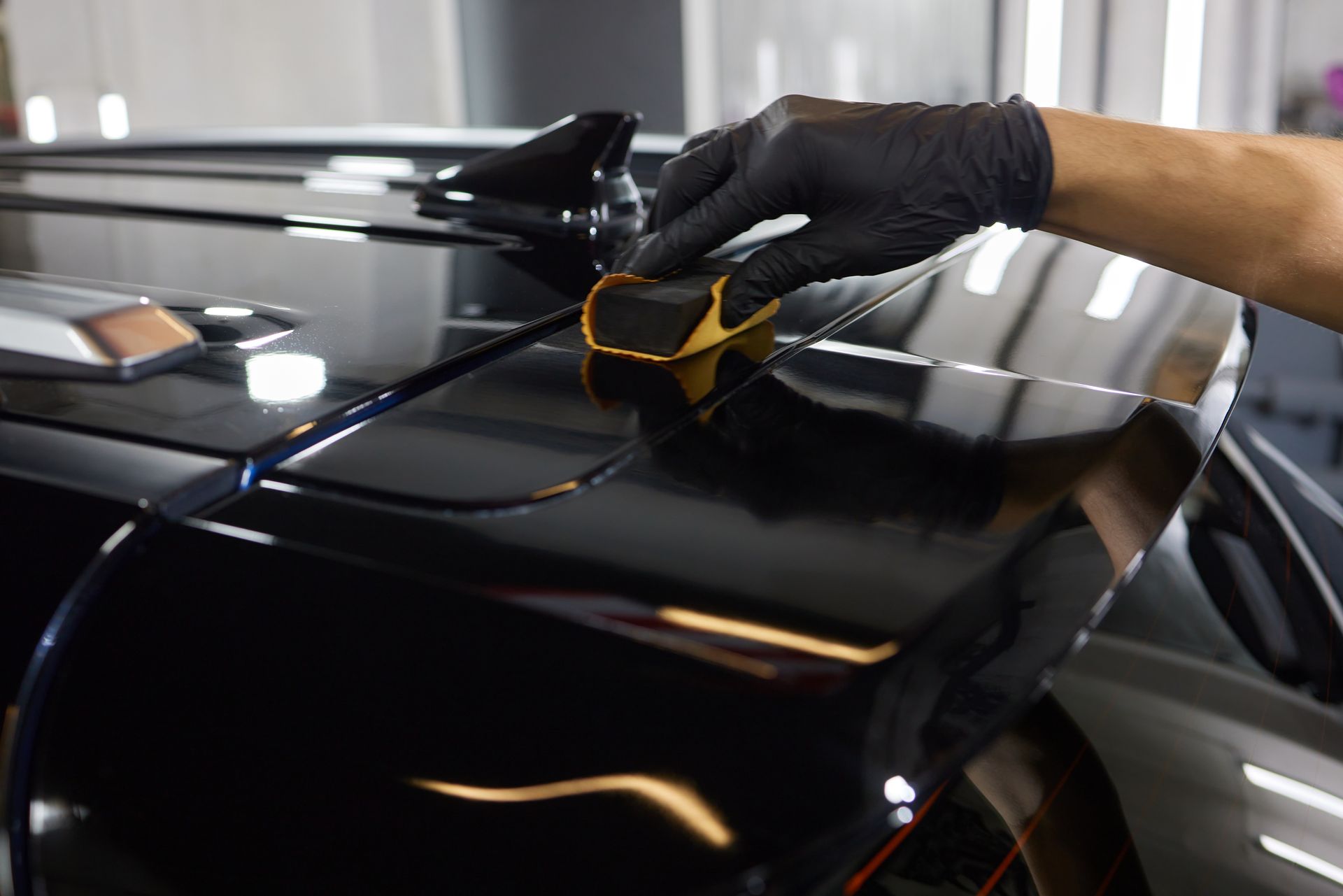 A person wearing black gloves applying a gold-colored coating to the black spoiler of a car.