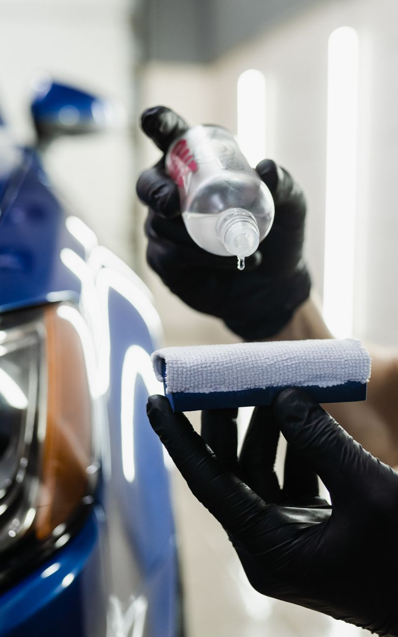 Gloved hands applying liquid from a bottle to a car with an applicator.