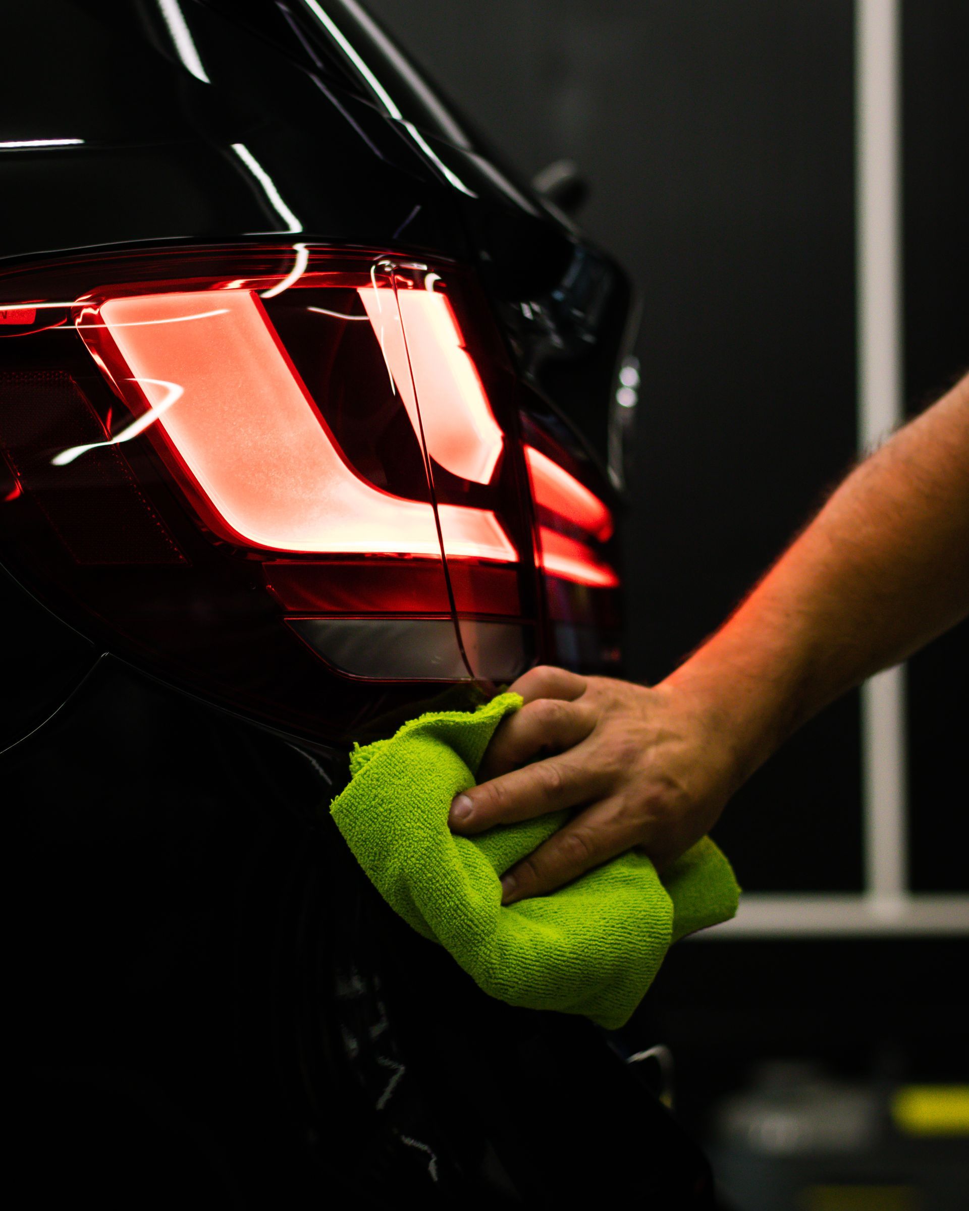 Person wiping the back of a black car with a green cloth; red taillights illuminated.