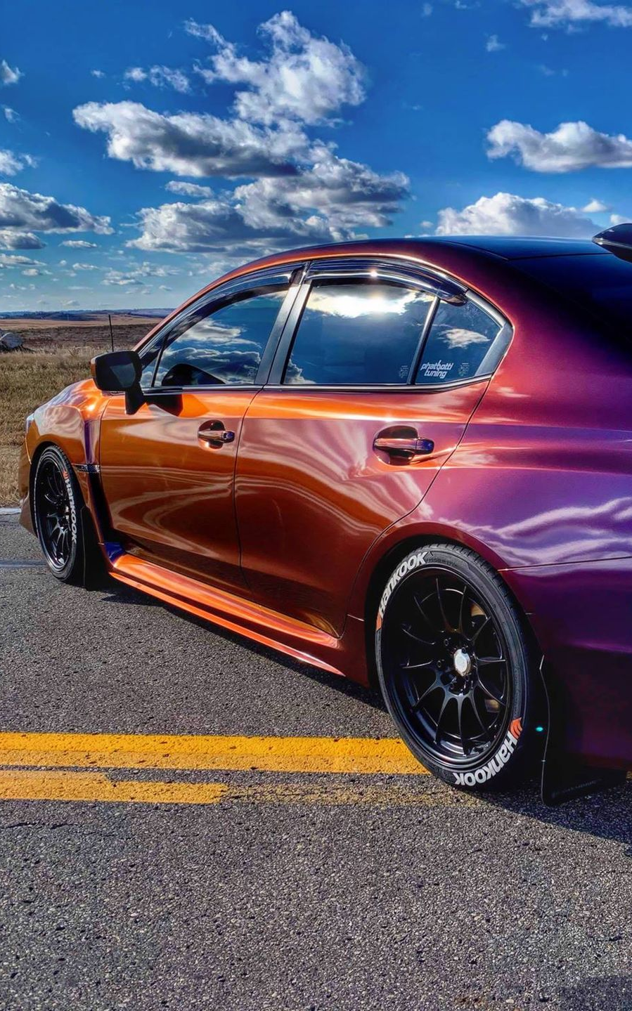 Custom painted orange and purple Subaru sedan on asphalt road, black wheels, against blue sky with clouds.