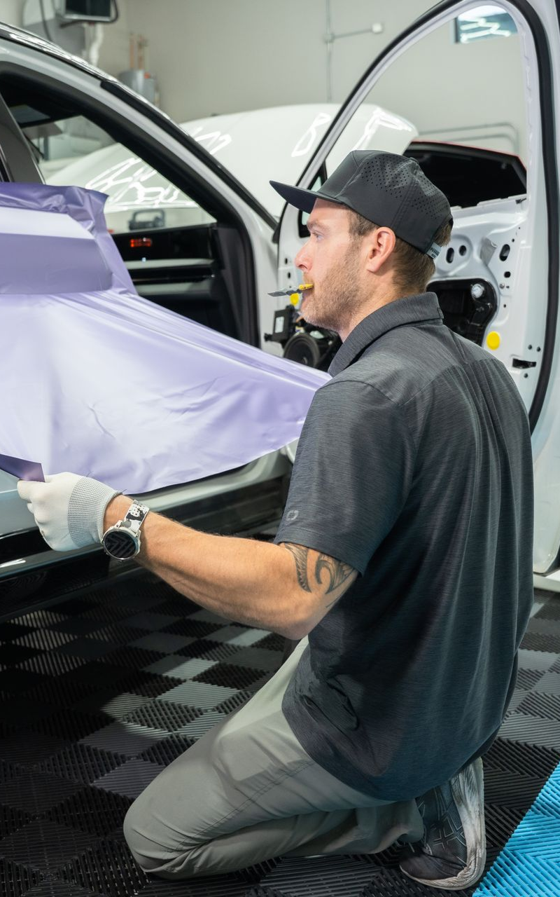 Man kneeling, applying purple wrap to a white car door in a garage, wearing a hat and gloves.