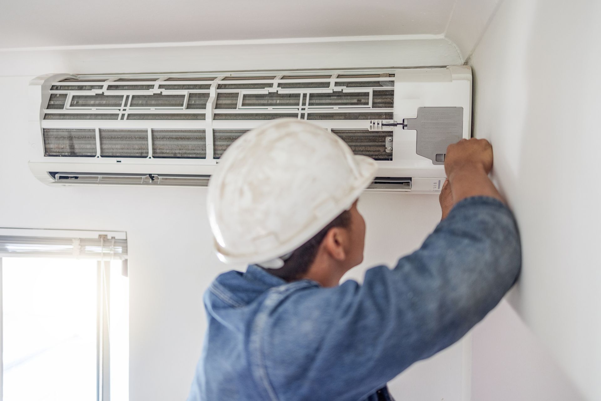 A man wearing a hard hat is fixing an air conditioner on the wall.