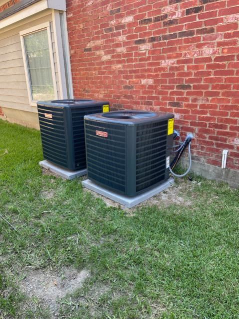 Two air conditioners are sitting in the grass in front of a brick building.