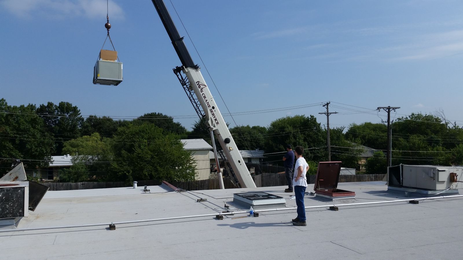 A man is standing on a roof watching a crane lift a piece of equipment.