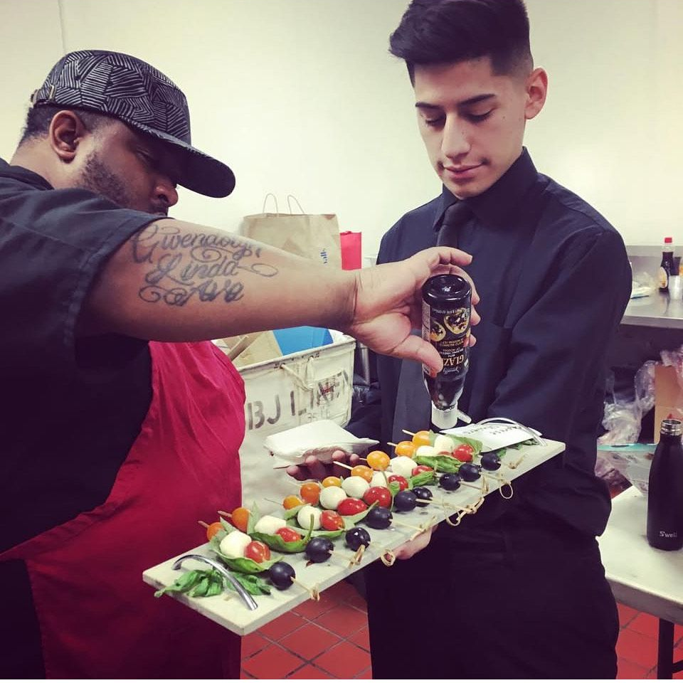 Two people arranging appetizers on a platter: chef drizzling sauce, server holding dish. Kitchen setting.
