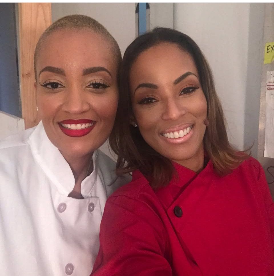 Two women in chef coats smiling, one with a shaved head and red lipstick, the other with brown hair and a red coat.