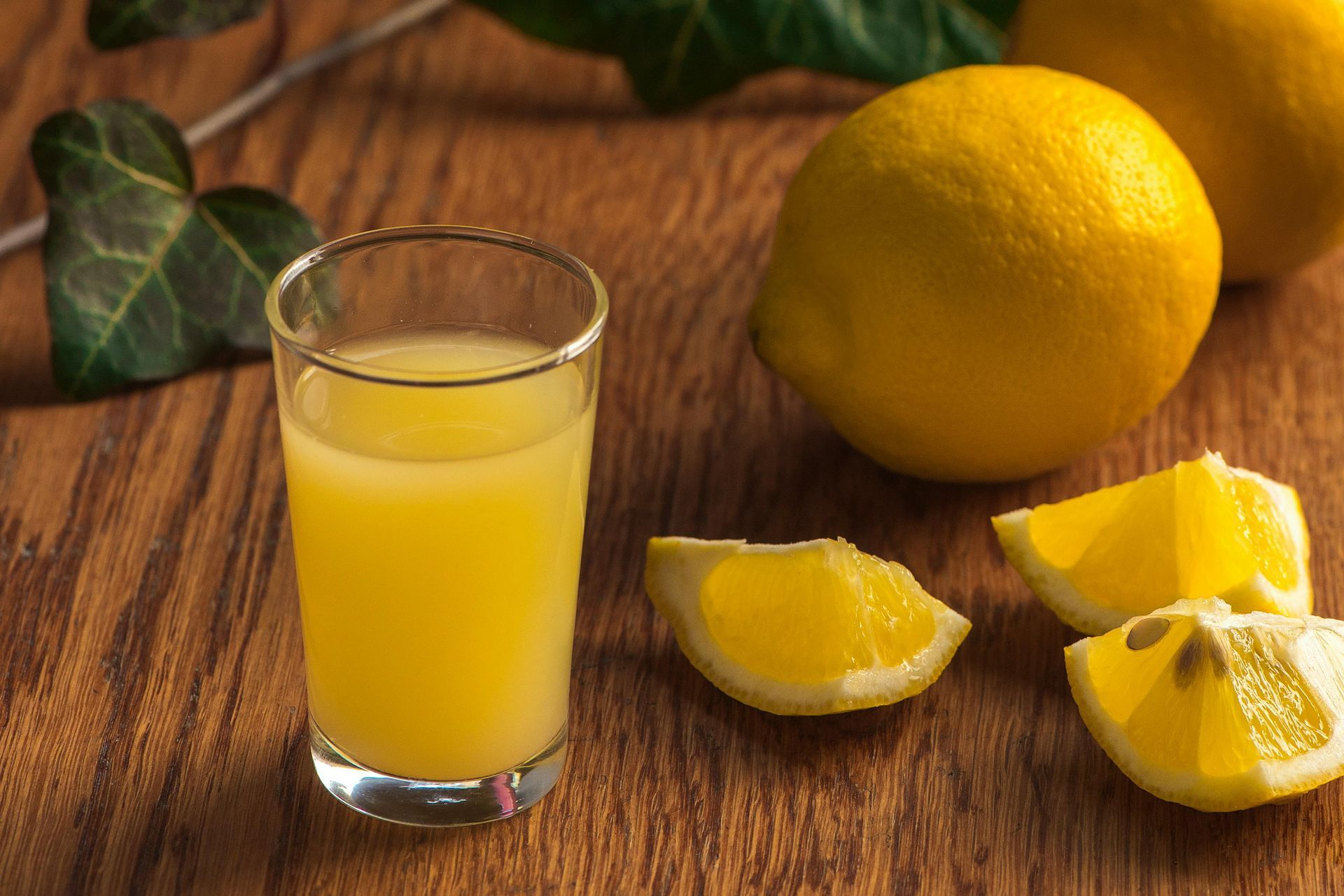 Glass of yellow lemon juice, lemons, and lemon wedges on wood.