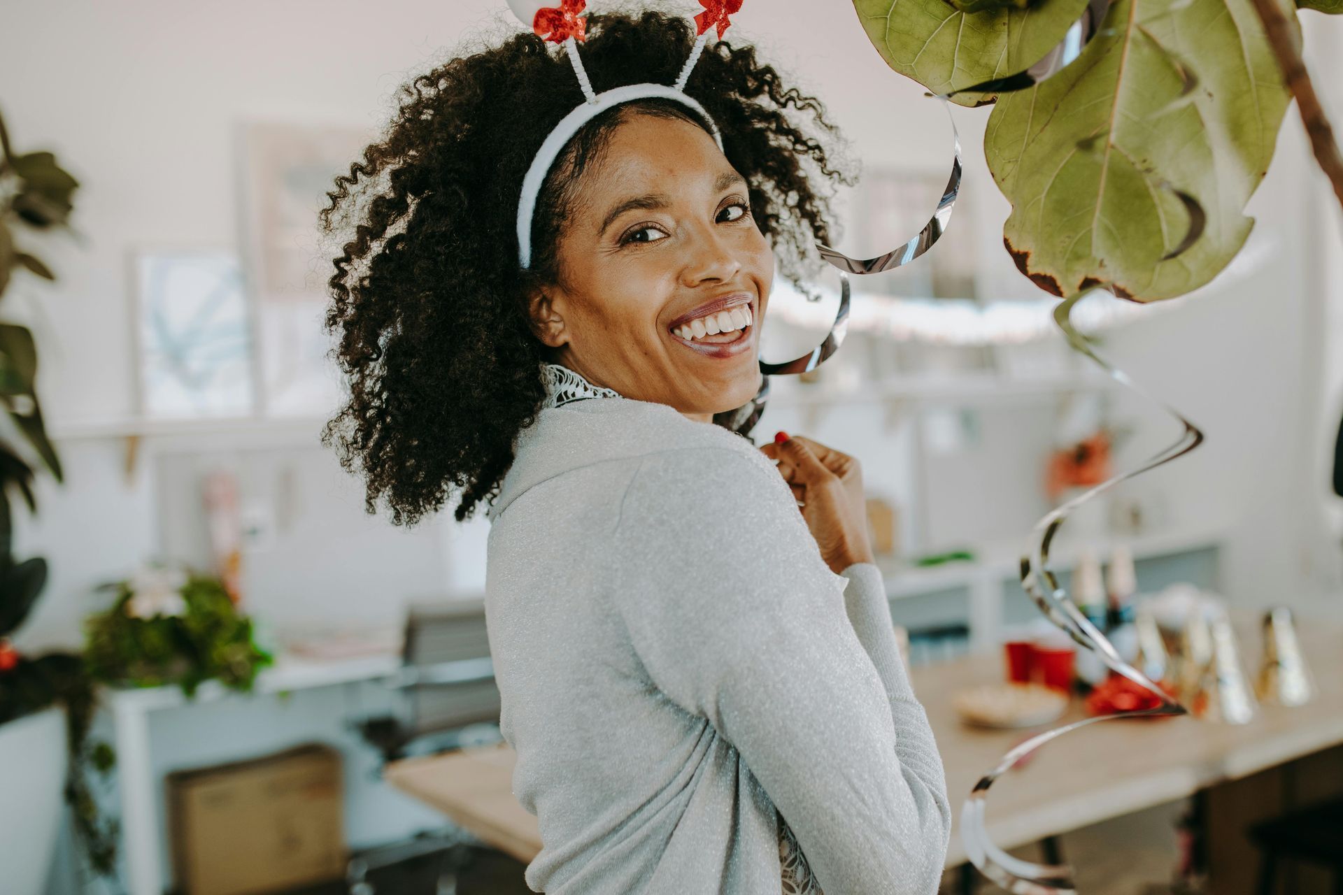 Woman with curly hair and reindeer antlers smiles, in a bright room with decorations.