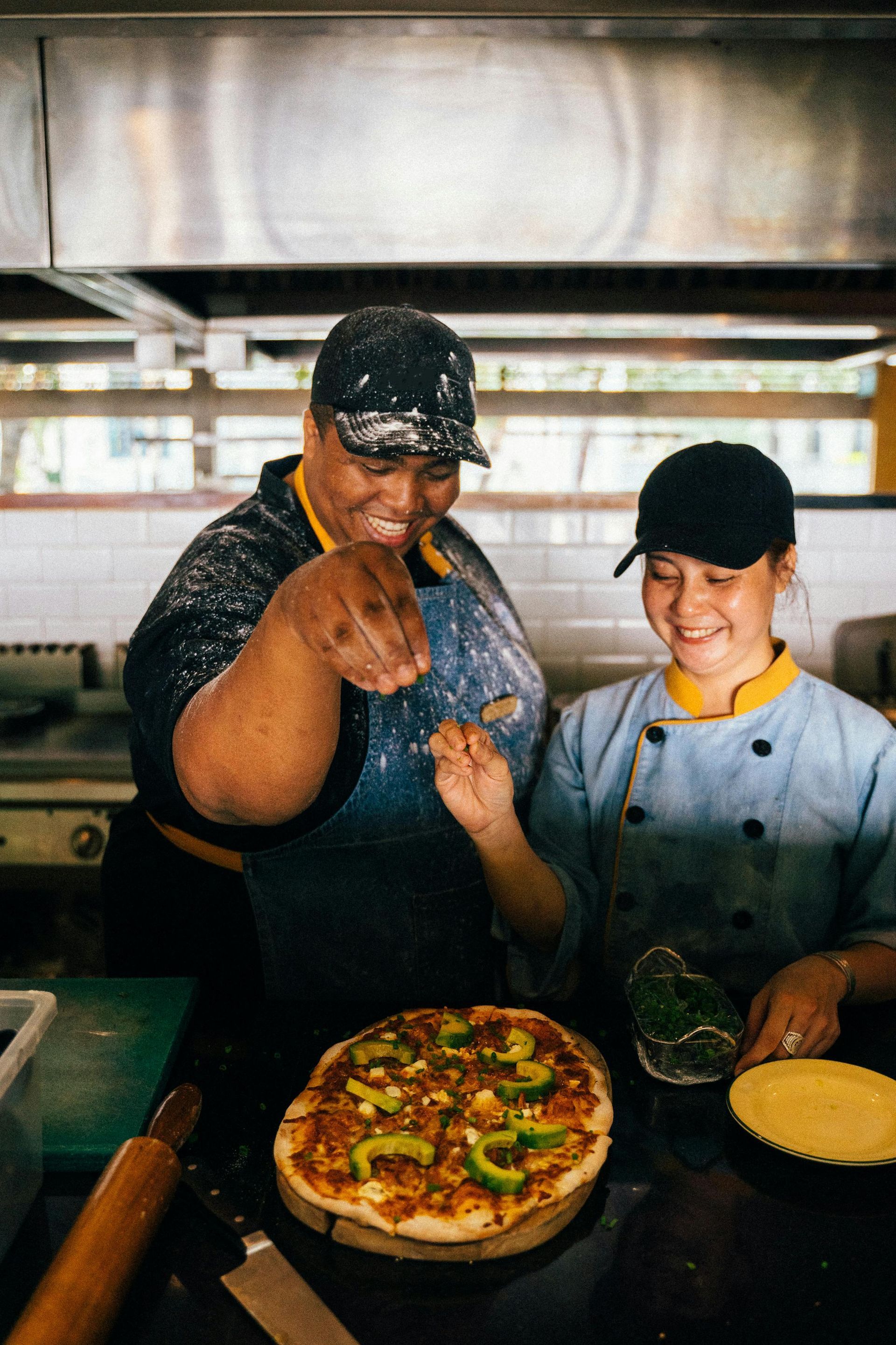 Two cooks preparing pizza in a commercial kitchen, sprinkling toppings, smiling. Pizza is on a wooden board.