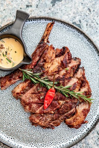 Sliced grilled steak with bone, sauce, rosemary, and chili pepper on a patterned plate.