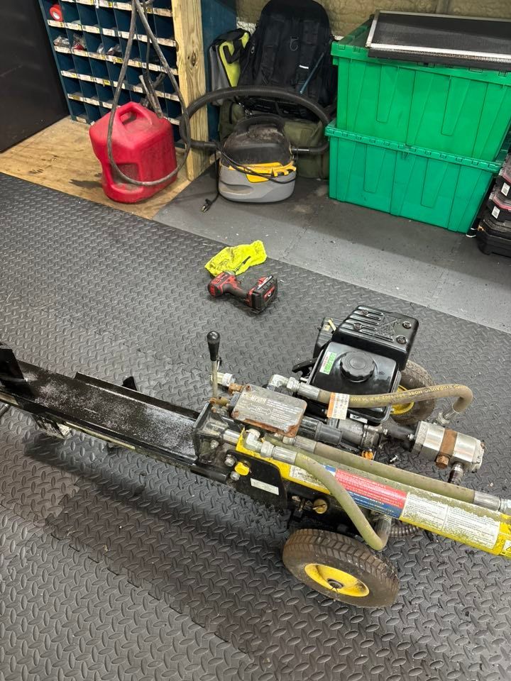 Log splitter on a black mat in a workshop with a gas can and green storage bins.