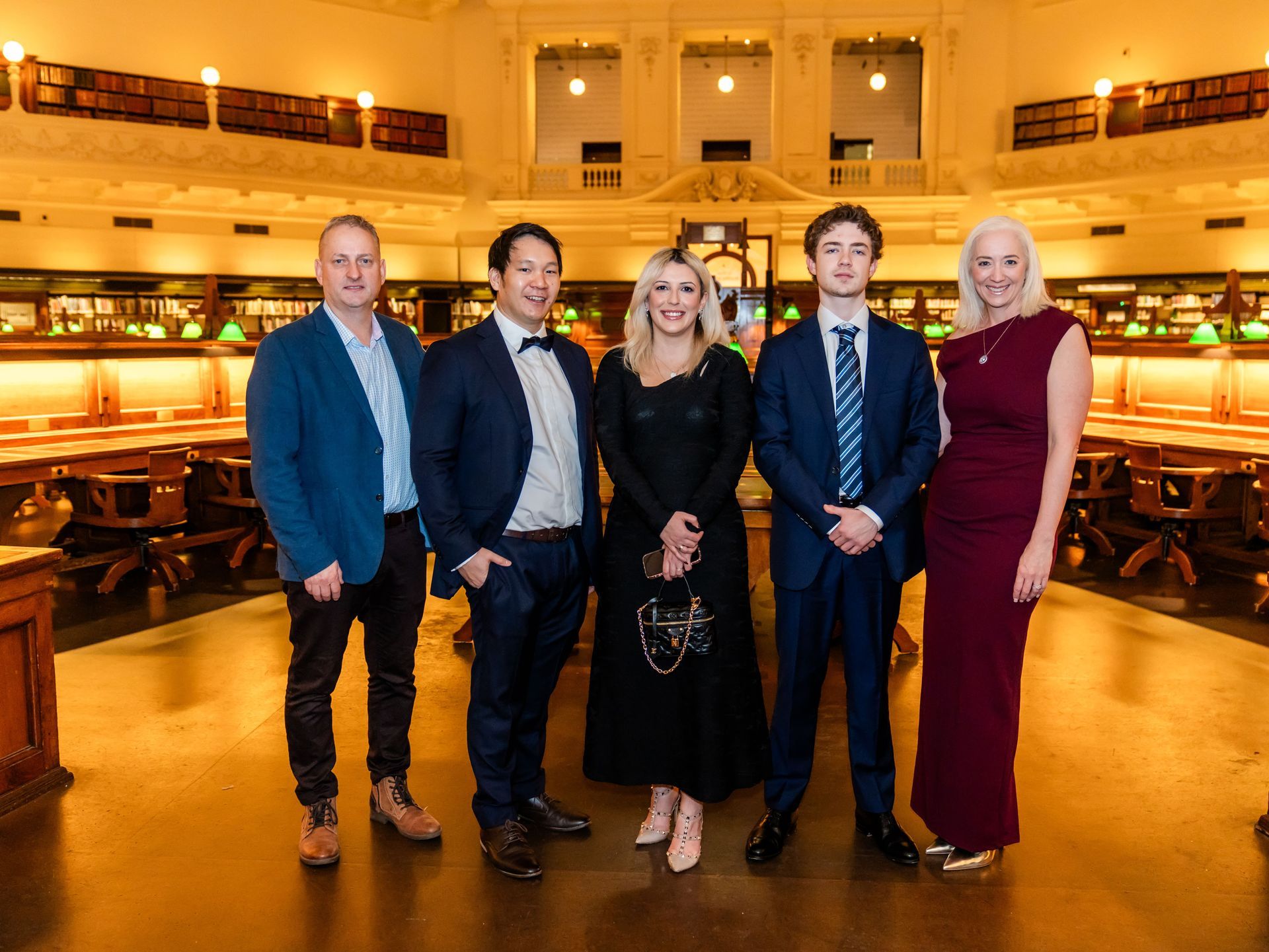 Five people posing in a warmly lit theater lobby with red velvet seating and balcony tiers