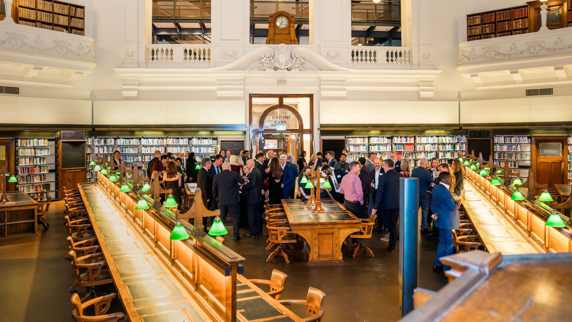 Busy library reading room with people around long wooden tables and green-shaded lamps