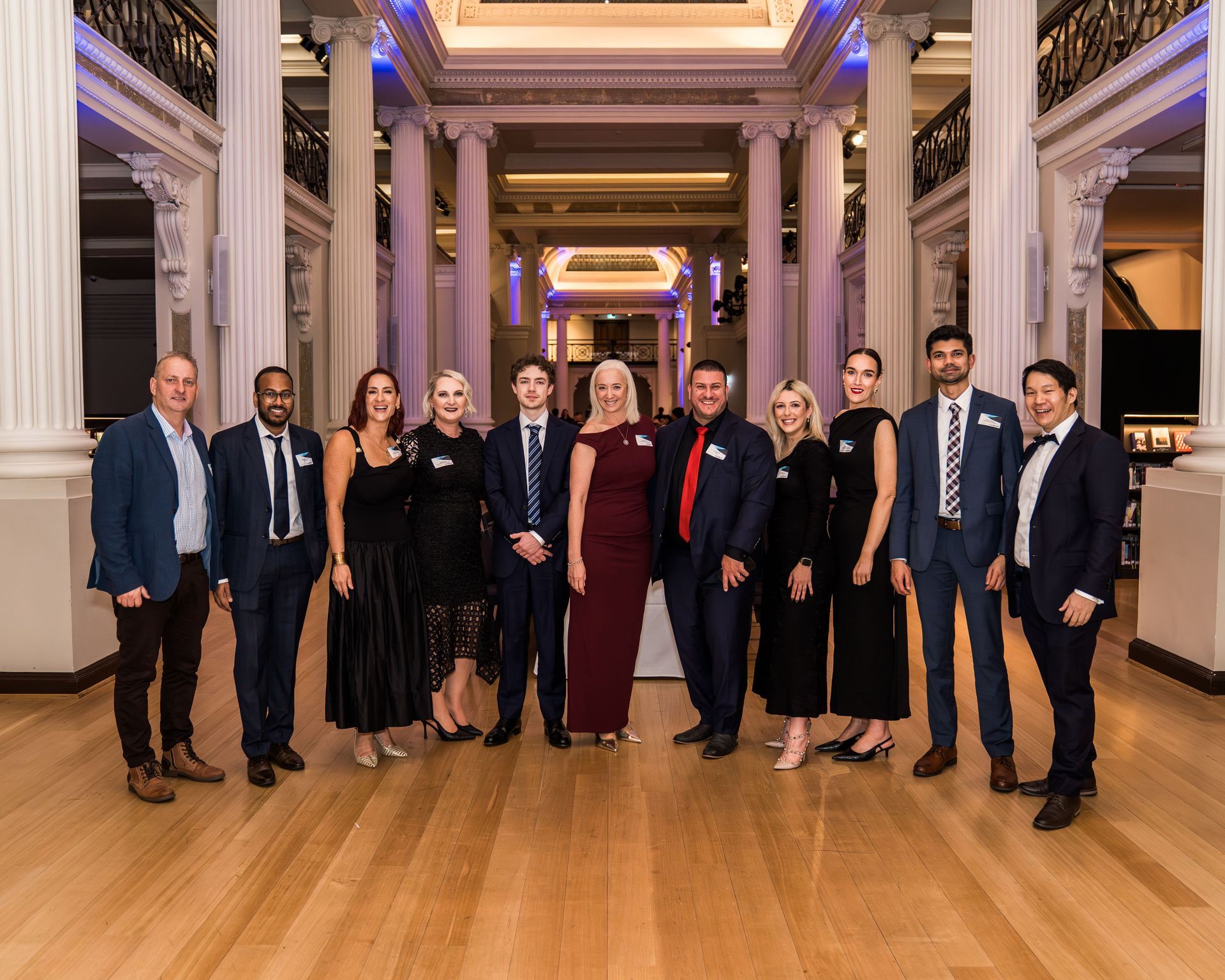 Group of people in formal attire posing in a grand hall with columns and chandeliers.