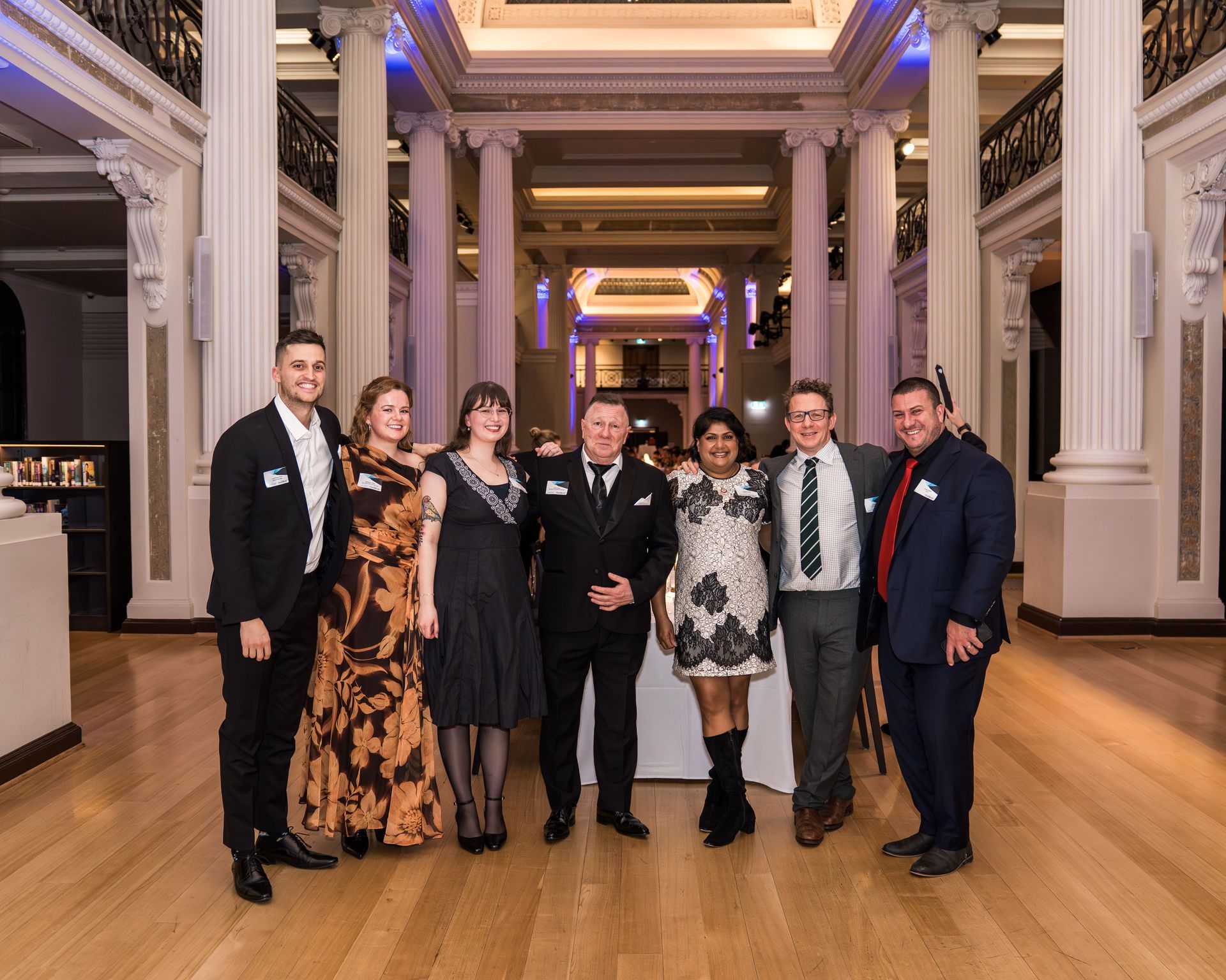 Seven people posing in formalwear in a grand ballroom with tall columns and polished wood floors.