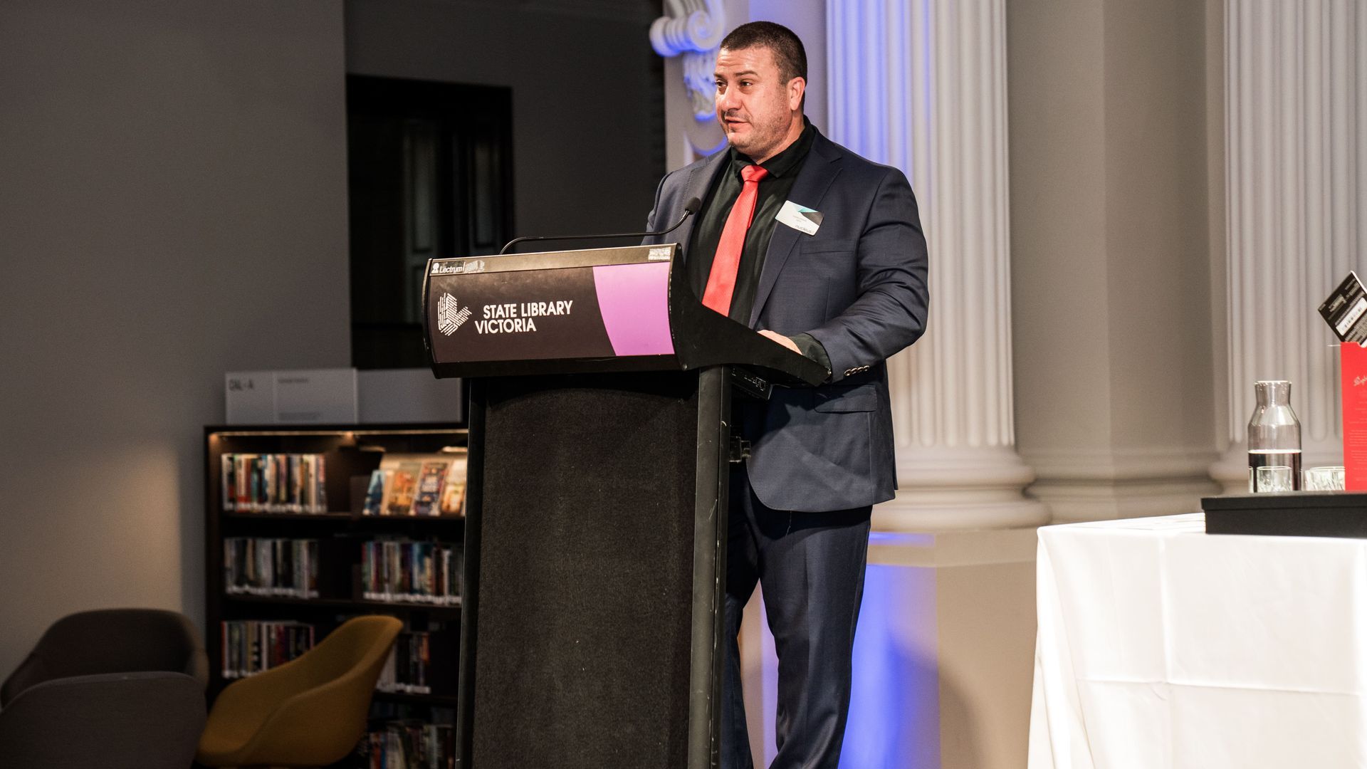 Man in a dark suit speaks at a podium in a formal room with columns and a lit screen behind him.