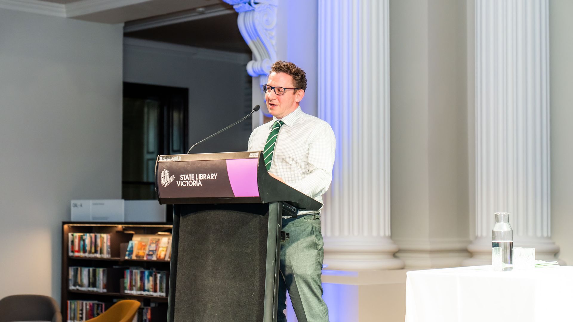 Speaker at a podium in a white shirt and green tie, addressing an audience in a formal hall