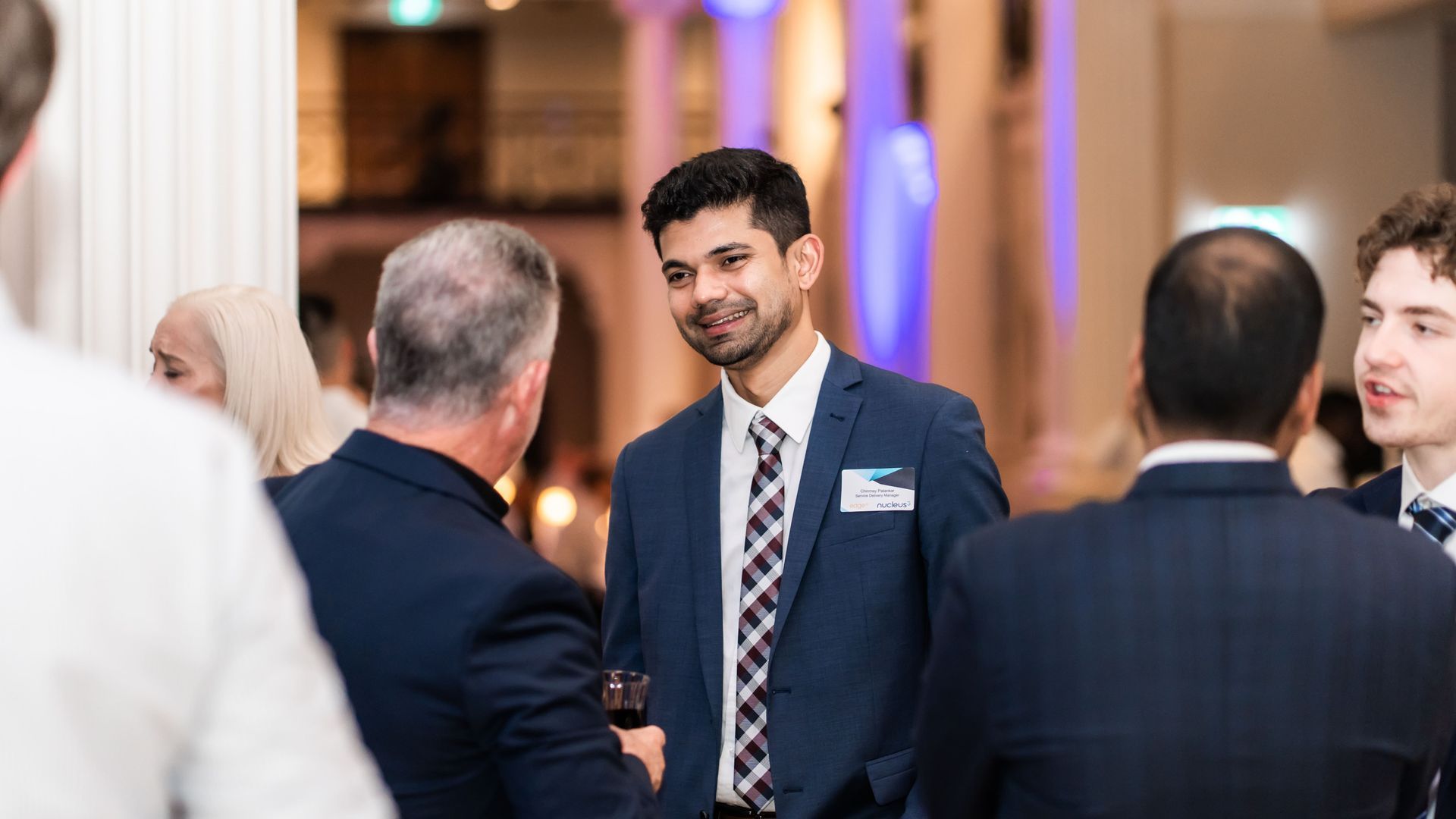 Man in a navy suit shaking hands at a formal indoor event with blurred guests nearby