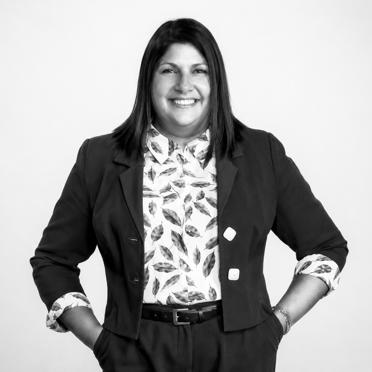 Smiling person in a dark blazer and patterned shirt, standing with hands in pockets on a white background