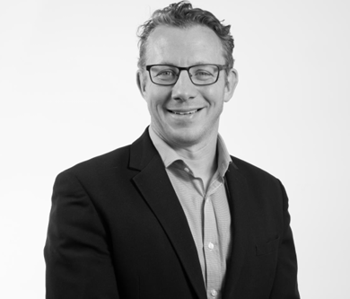 Smiling man in glasses wearing a dark blazer over a light shirt against a plain background