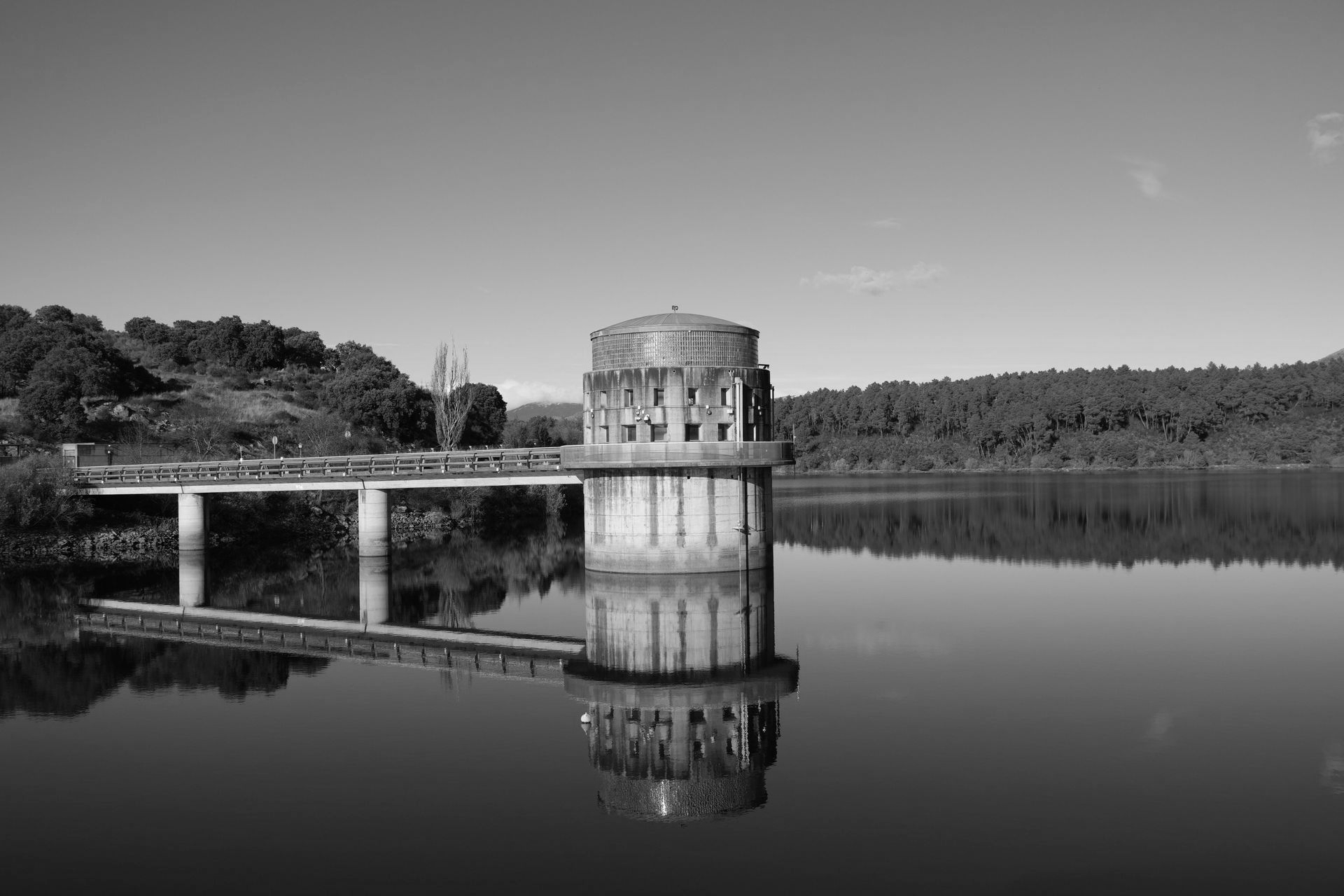 Black-and-white view of a small stone tower beside a bridge, reflected in a calm lake