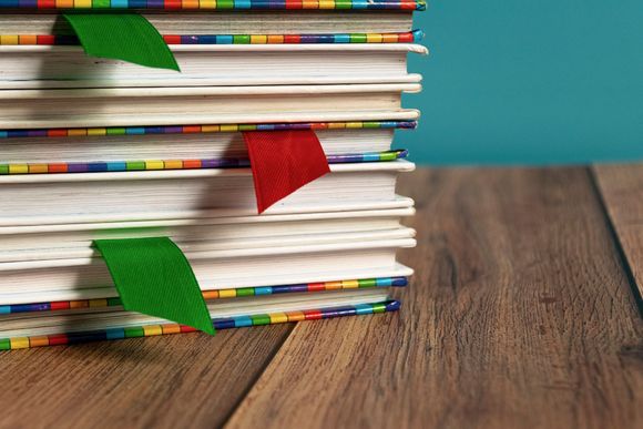 Pila di libri su una superficie di legno, con bordi colorati e segnalibri rossi e verdi.
