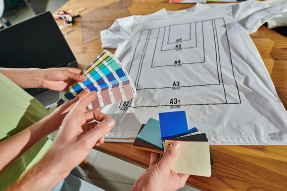 Mani che tengono campioni di colore e il layout del disegno su una maglietta bianca, mentre lavorano su un disegno di stampa.