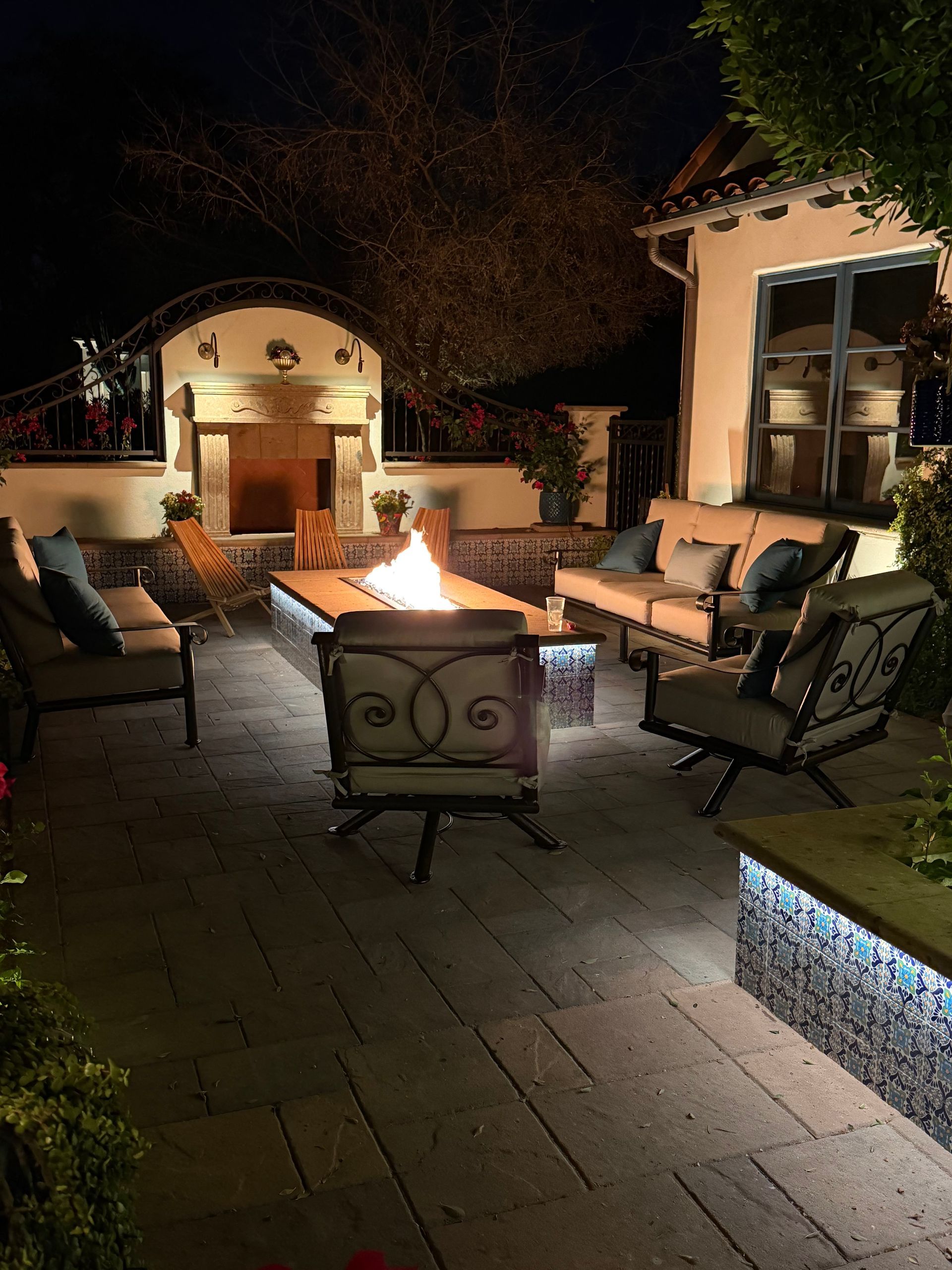 Outdoor patio with cushioned chairs and a fire pit at night, lit by warm lights and blue pathway lighting