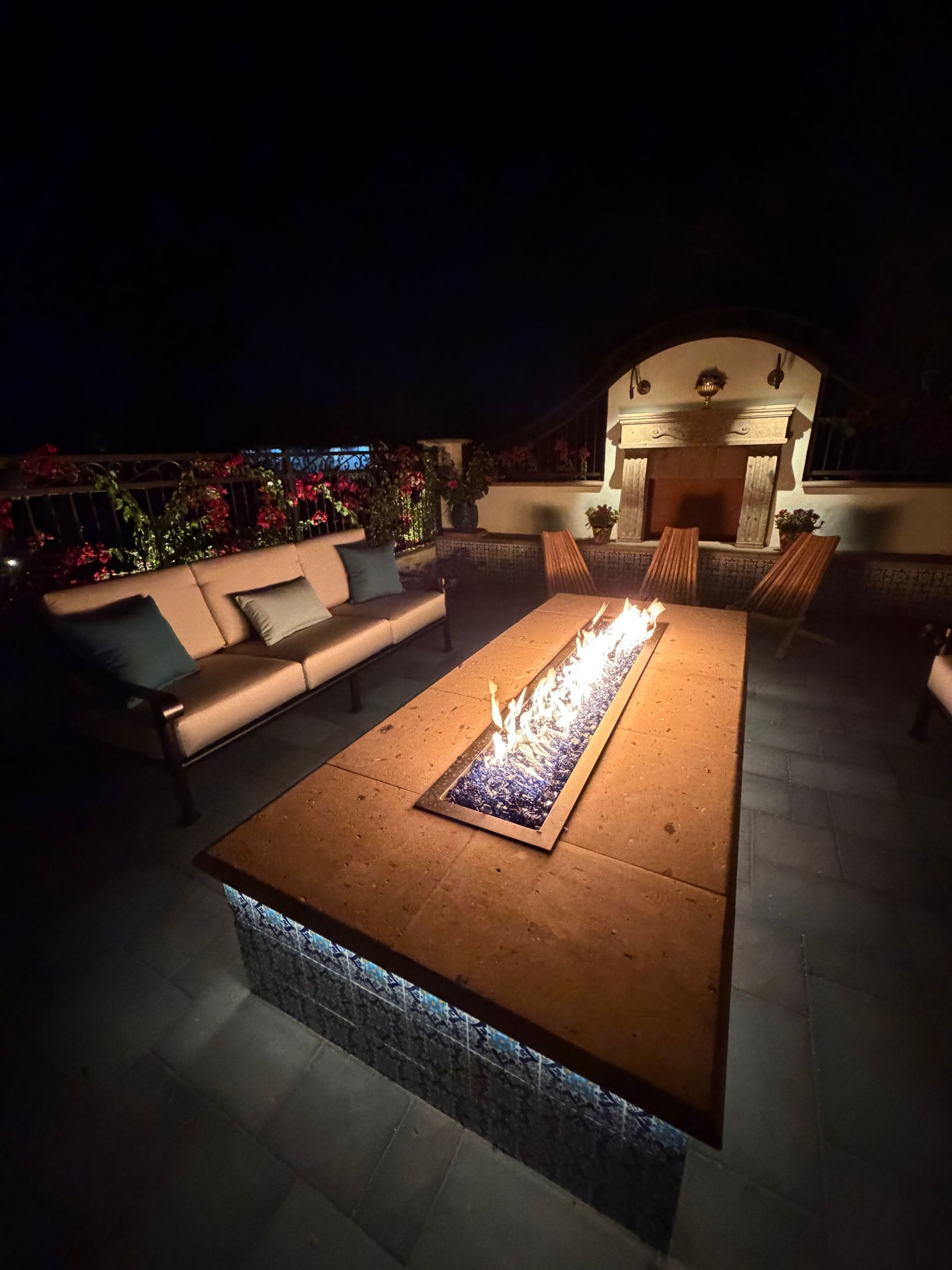 Modern patio at night with a long fire pit, cushioned sofa, and lit outdoor fireplace