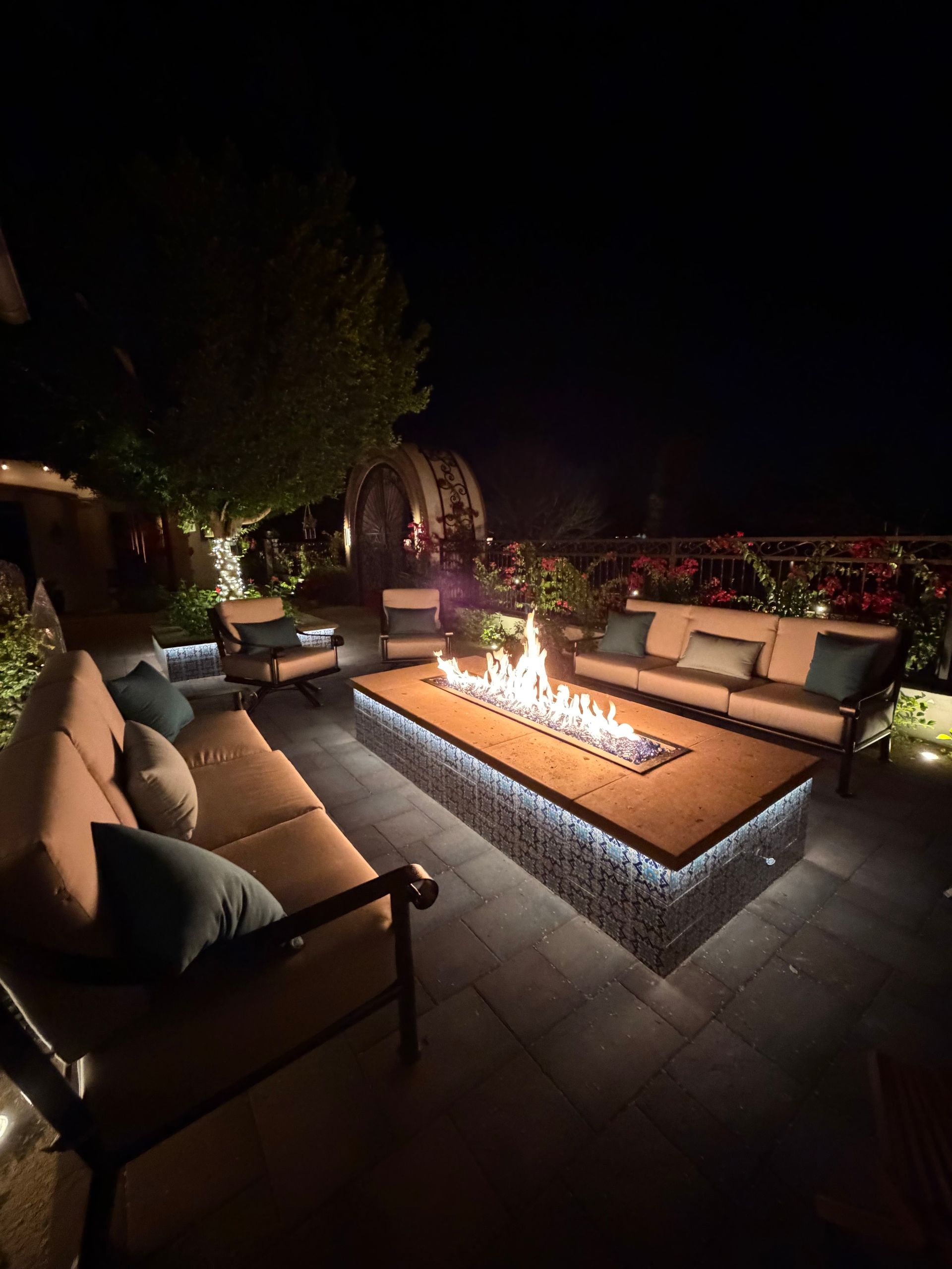 Outdoor patio with cushioned seating around a rectangular fire pit at night, lit by warm flames.