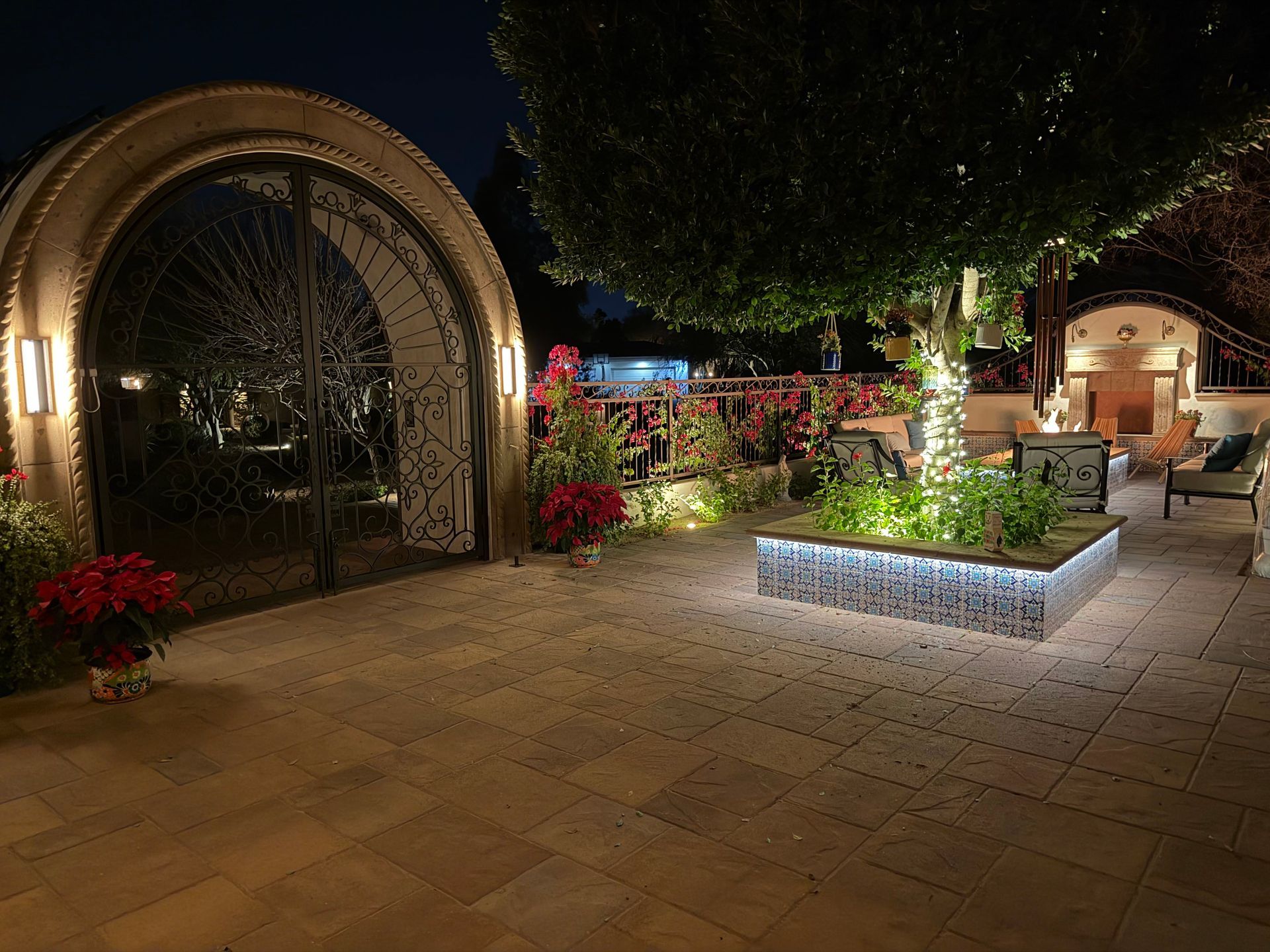 Nighttime patio with arched gazebo, lit planters, and flowering plants under trees