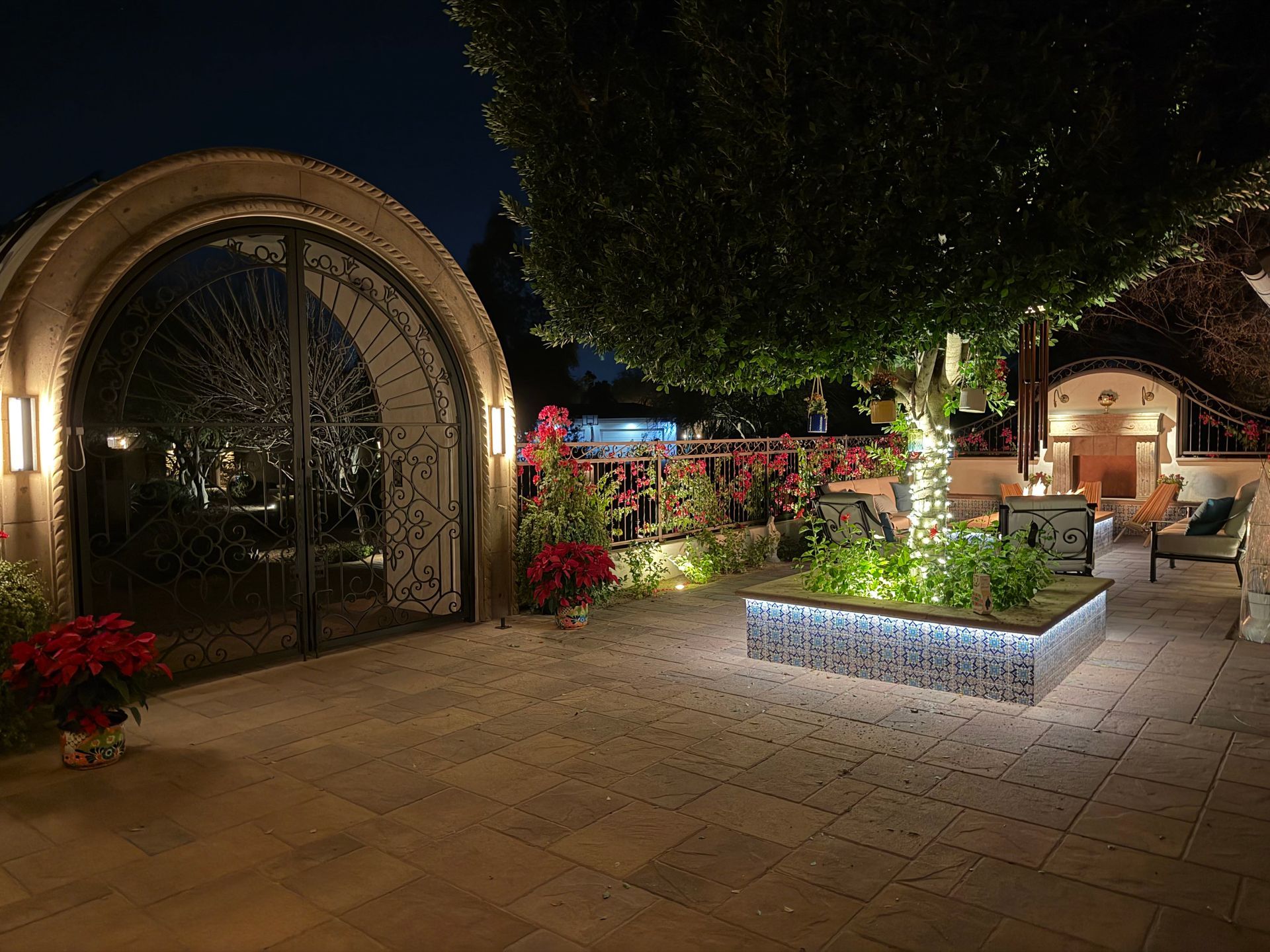 Nighttime garden courtyard with arched entrance, lit planters, and stone patio chairs