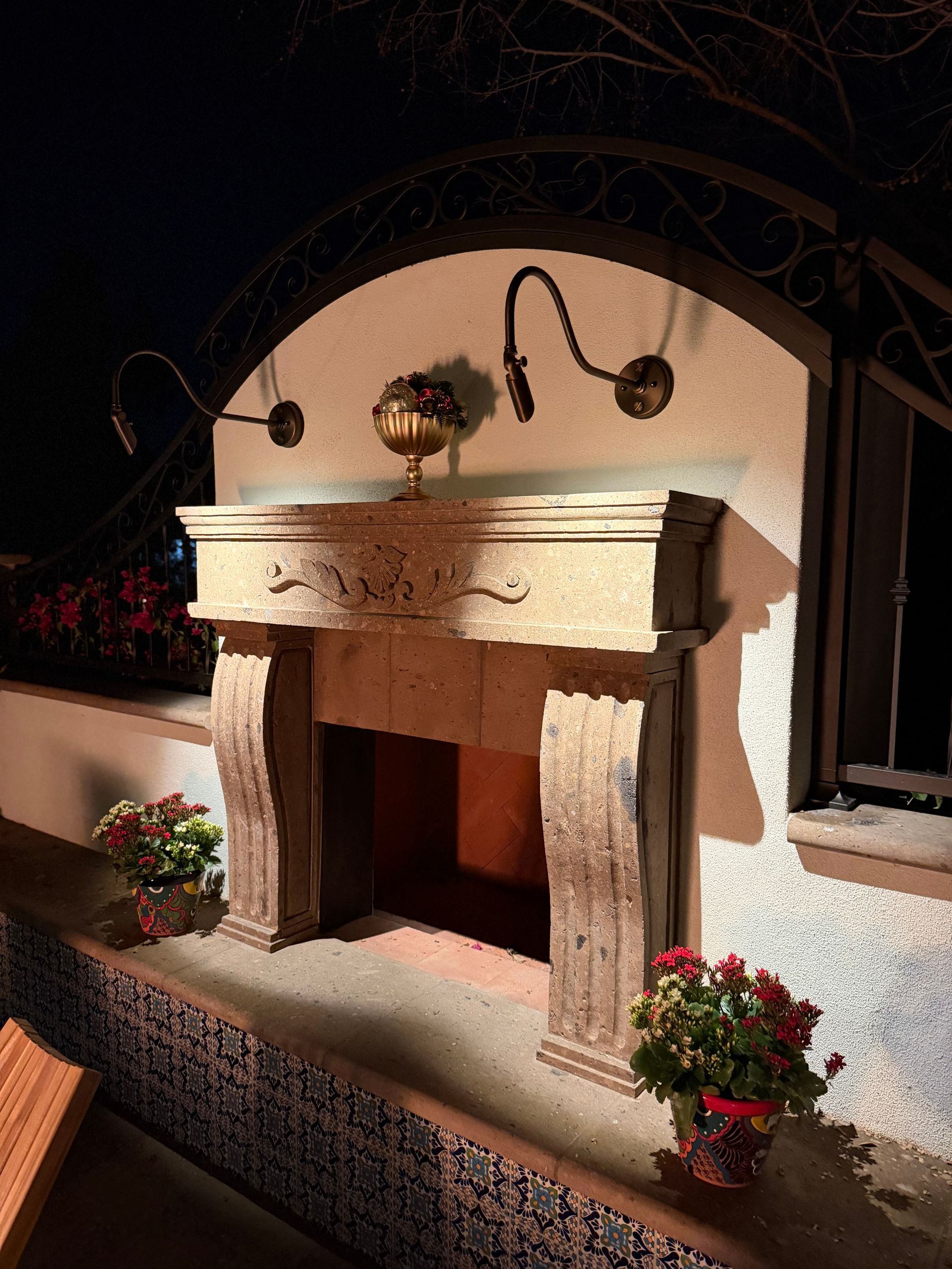Decorative outdoor fireplace with stone mantel, wall lamps, and potted flowers at night.