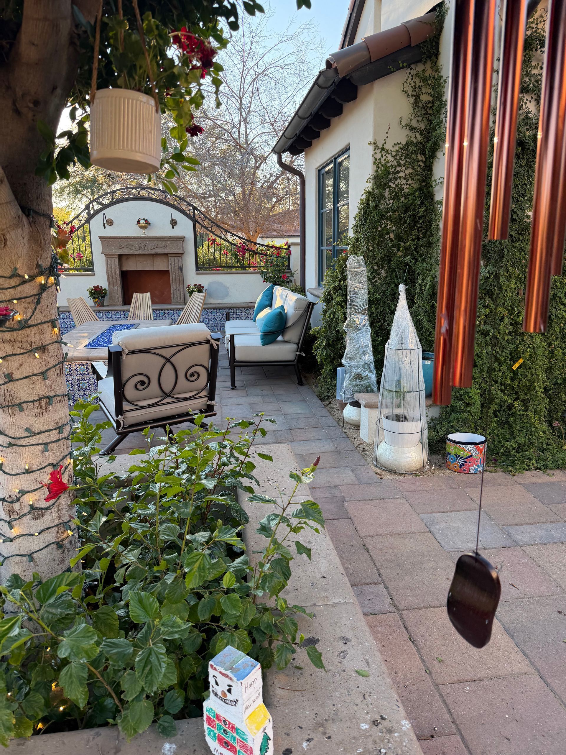 Patio garden with stone path, cushioned seating, hanging lanterns, and potted plants beside a white wall