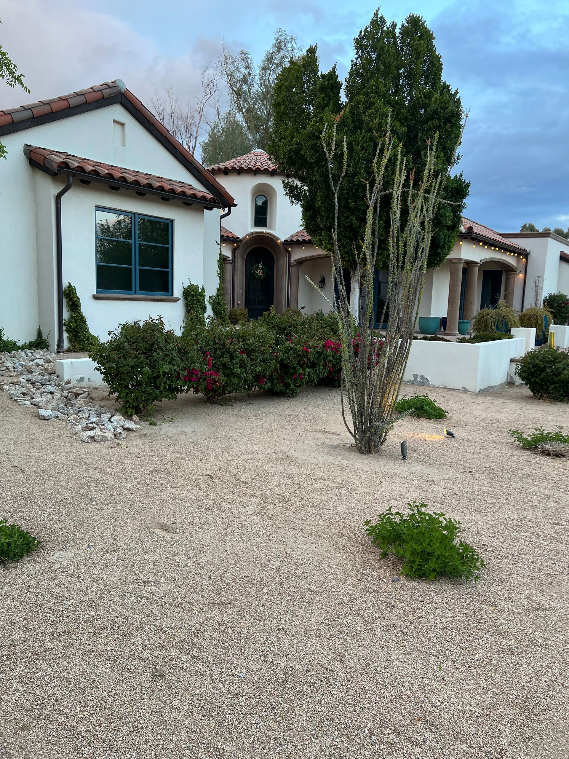 White stucco house with red tile roof, gravel yard, shrubs, and a tall tree by the entrance