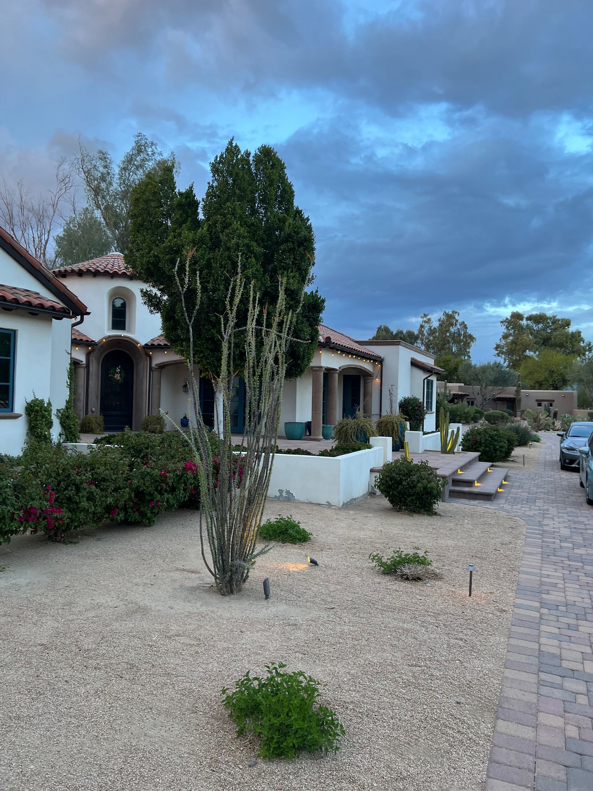 White stucco house with arched doorway, gravel yard, and tall trees under a cloudy sky.