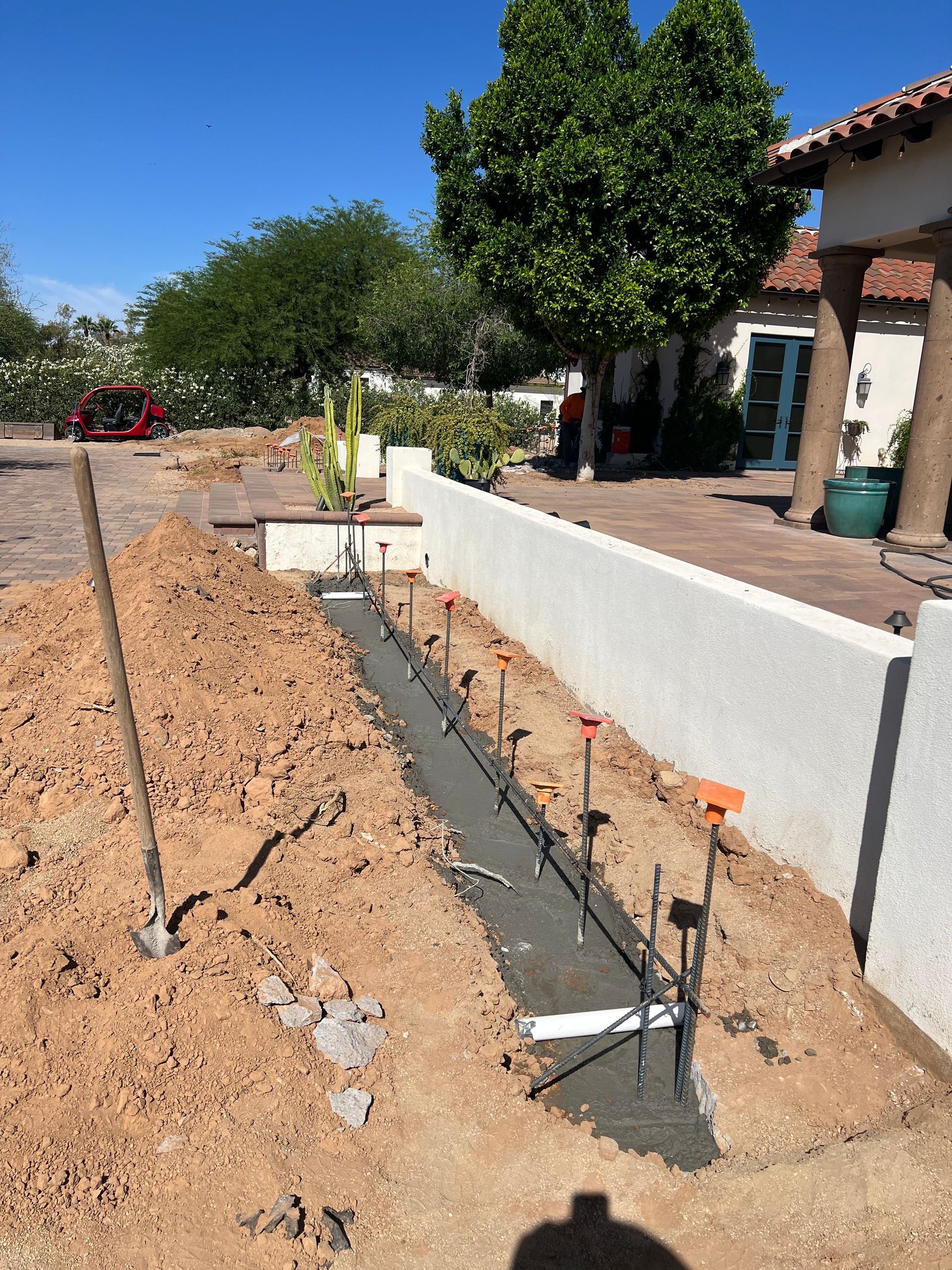 Outdoor construction site with trench, exposed rebar, and white wall beside a house on a sunny day