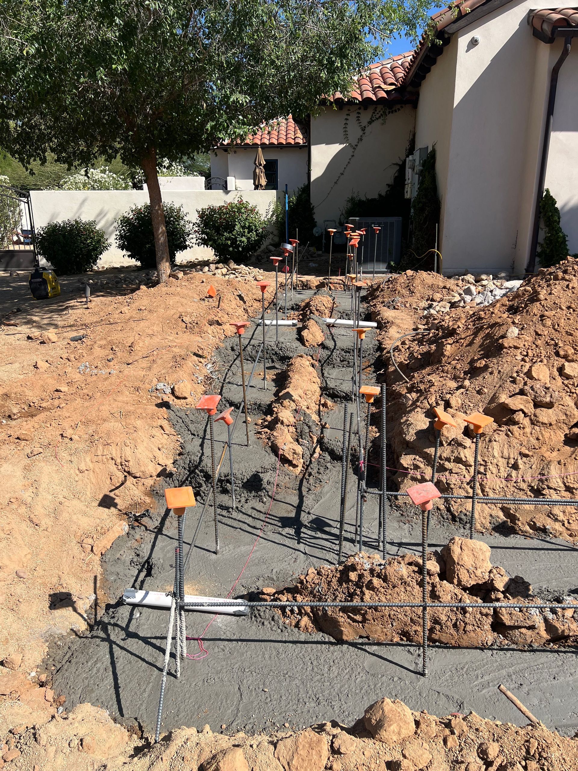 Foundation rebar and concrete footings in a dug trench beside a stucco house and tree-lined yard
