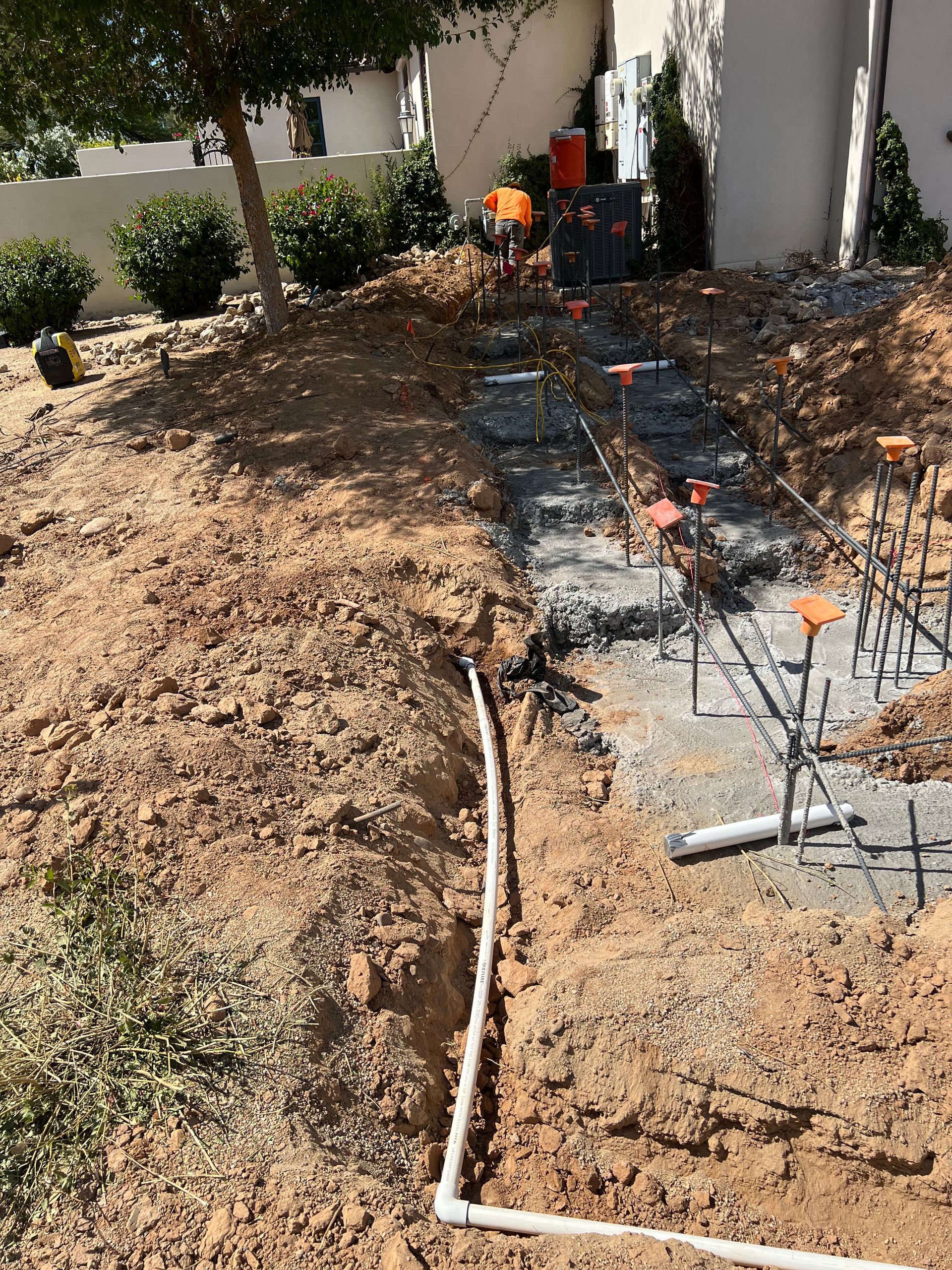 Construction site with trenching, exposed pipes, and workers in an outdoor yard beside a building