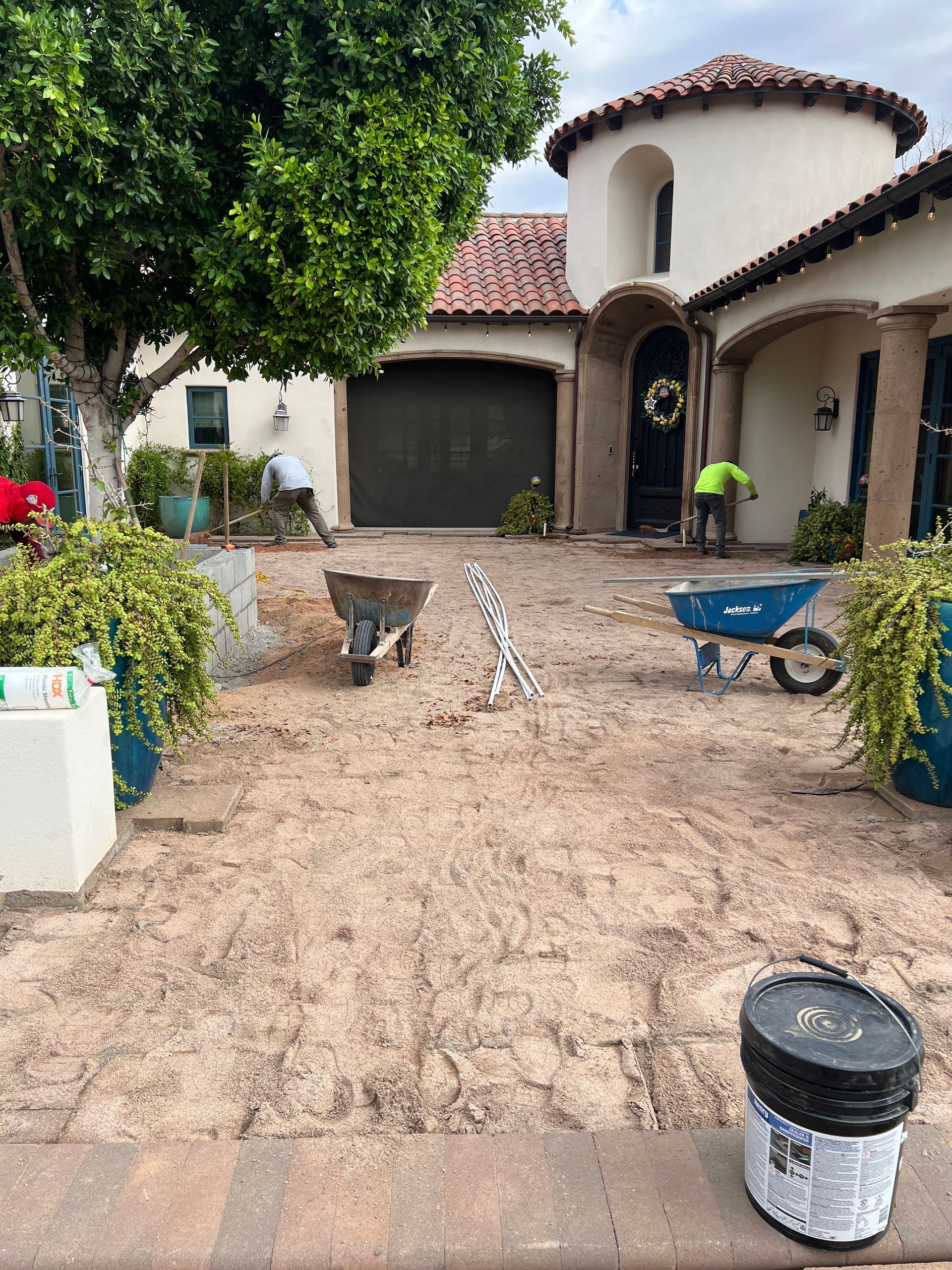 Front yard with wheelbarrows and a paint bucket outside a stucco house with a garage.