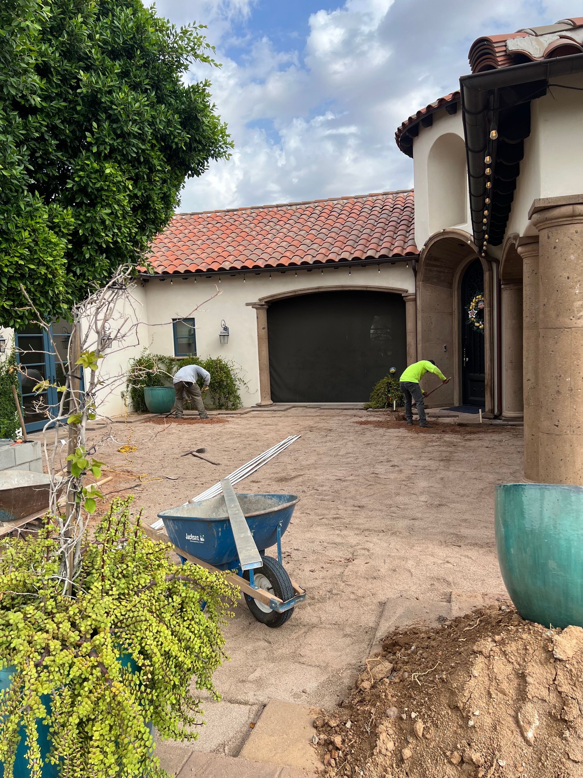 Wheelbarrow and dirt pile in a courtyard beside a stucco house and garage, with a person near the wall.