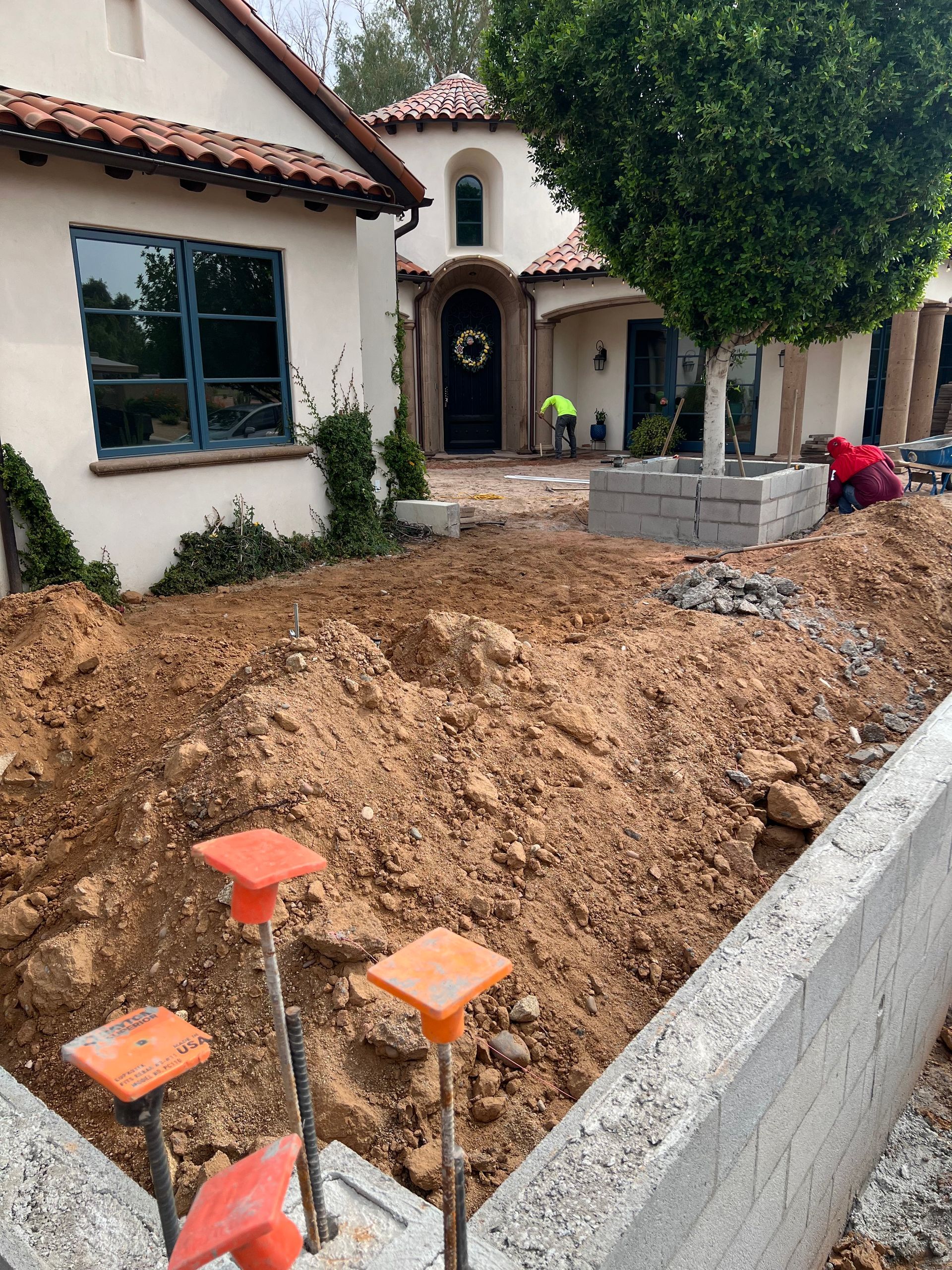 Front yard construction with dug-up soil, concrete curb, and a stucco house with arched entryway.
