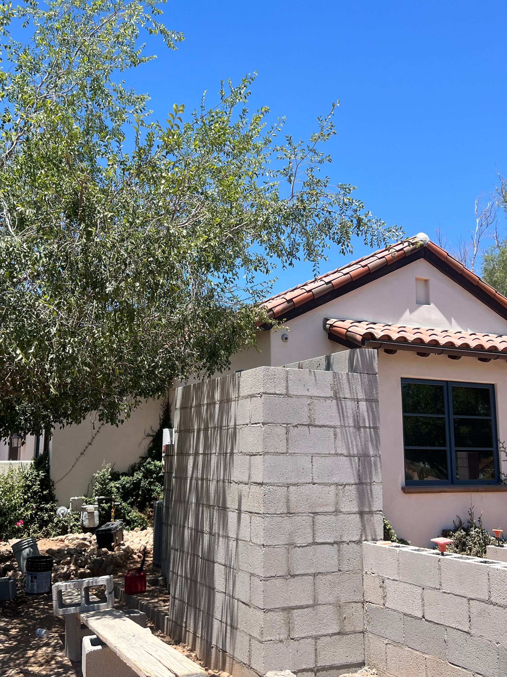 Sunlit house exterior with stucco walls, red tile roof, and a stone corner wall beside a tree